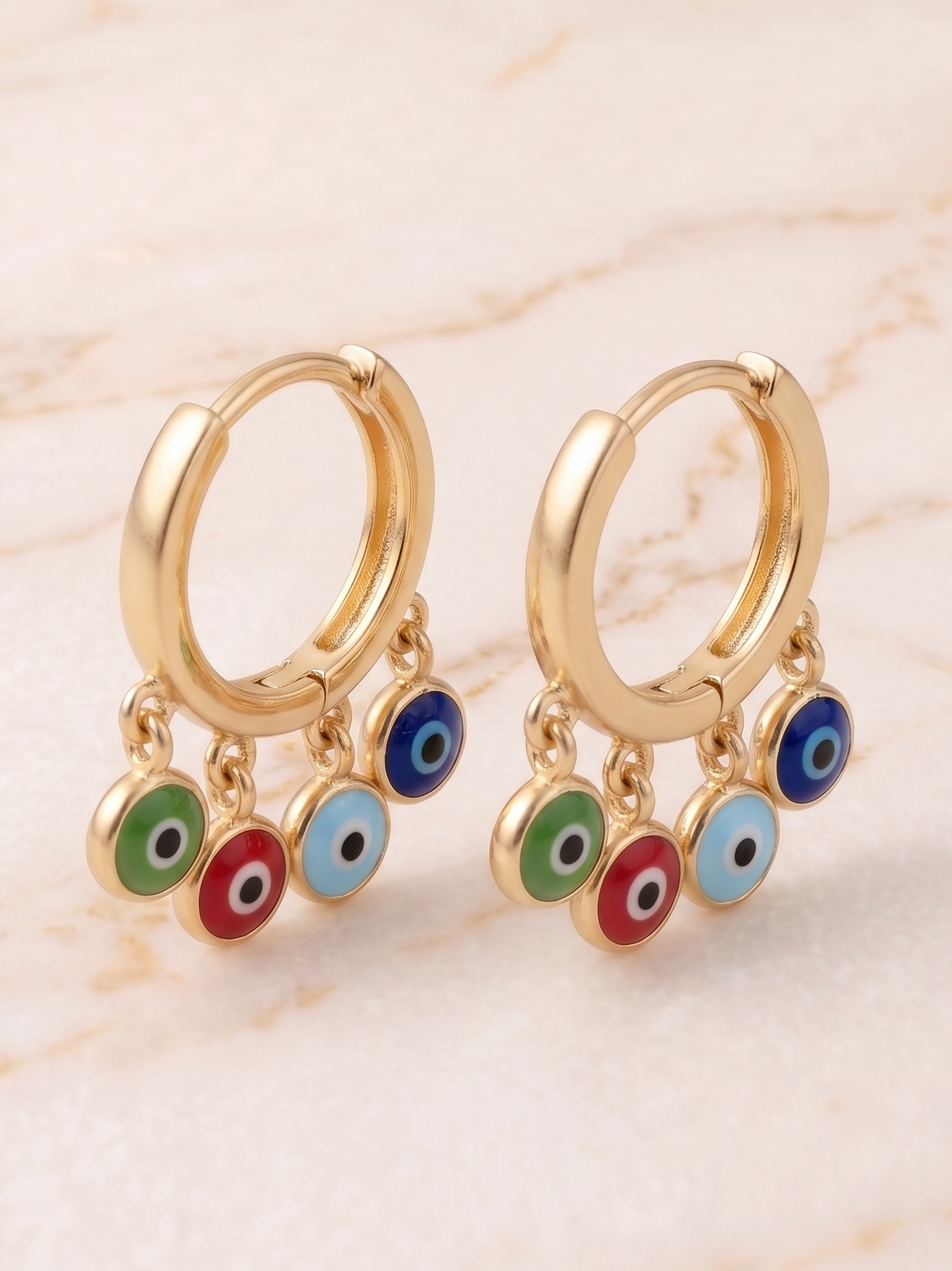 Gold hoop earrings with colorful evil eye charms on a beige marble background